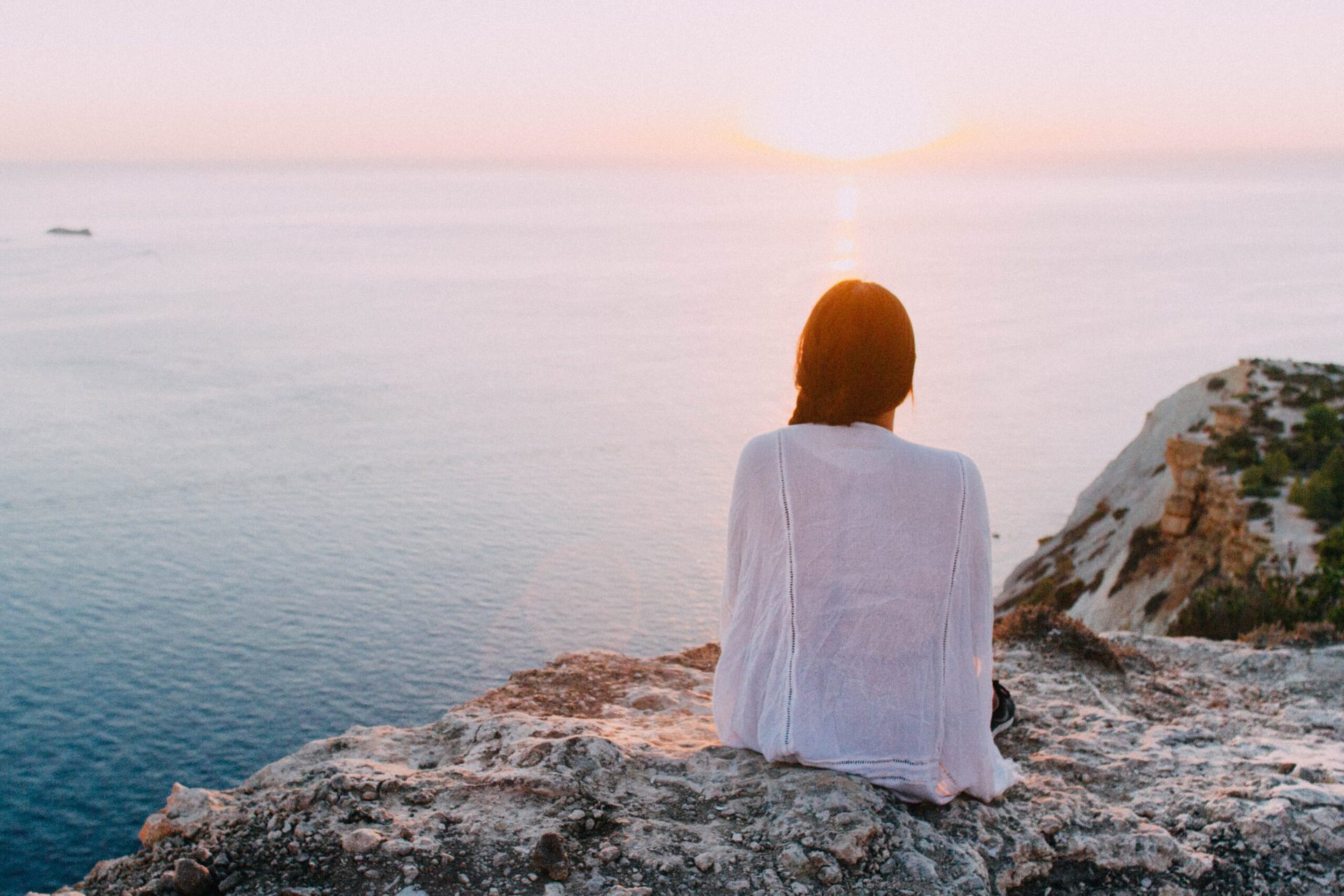 A woman in a pale top sits alone on a rocky cliff at sunset, looking out to the sea.