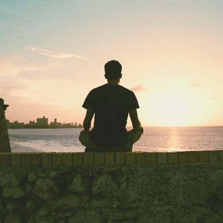 A man sits cross-legged on a stone wall at dusk, silhouetted against the sea and sky.