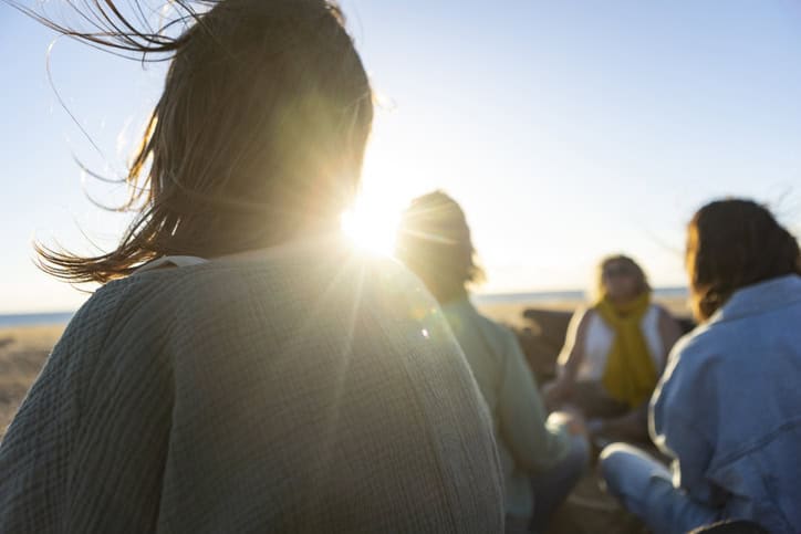 A small group gathered close together on a beach at sunset, backlit by low golden light.